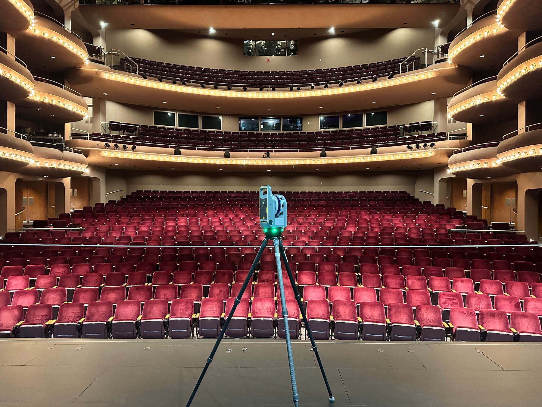 A Trimble X9 laser scanner on the stage of a performing arts venue, facing the empty auditorium and its tiered balconies.