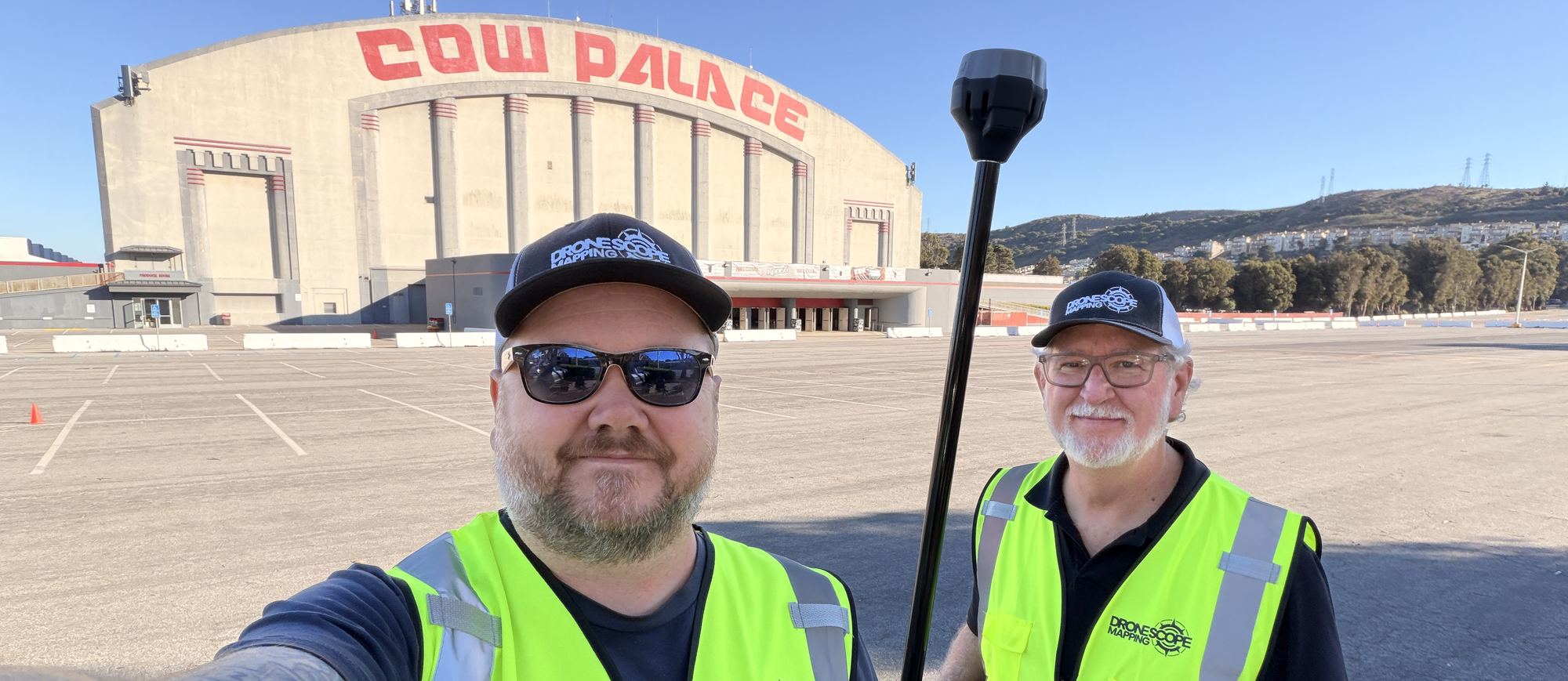 Two Drone Scope Mapping staff holding a GNSS rover pole in the parking lot of the Cow Palace arena, with the building and its signage behind them.