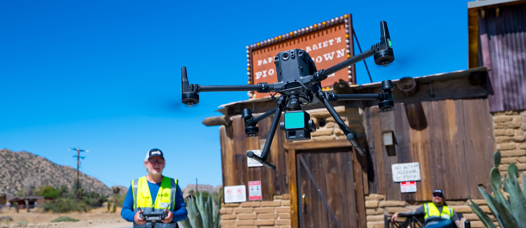 A DJI Matrice 350 RTK carrying an L2 LiDAR payload, hovering in front of a desert venue while a pilot in a high-visibility vest flies it from the ground.