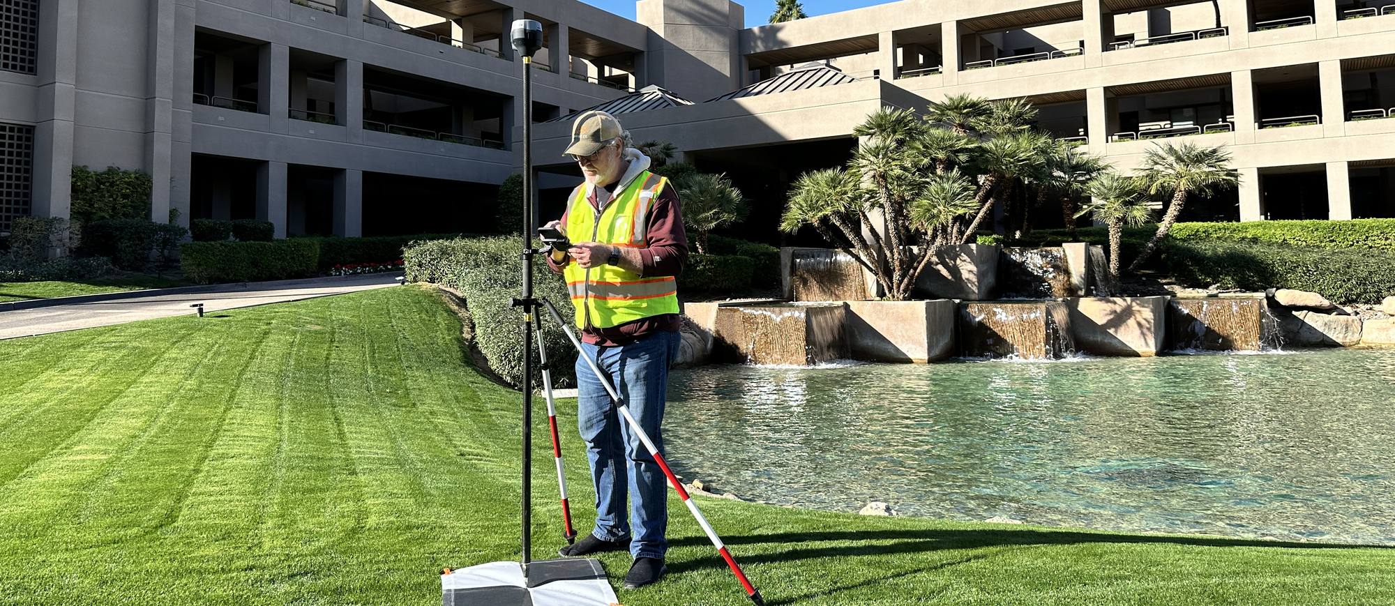A Drone Scope Mapping operator with a GNSS rover on the lawn of a large resort building, beside a landscaped water feature.