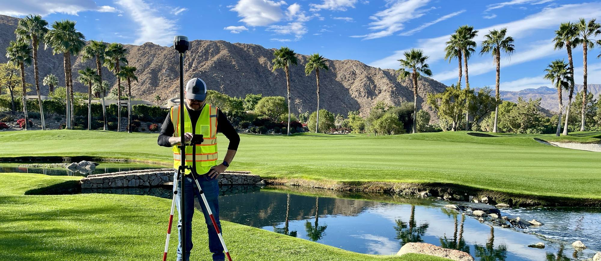 A Drone Scope Mapping operator setting a GNSS rover on the fairway of a desert golf resort, with palms, a water hazard and mountains behind.