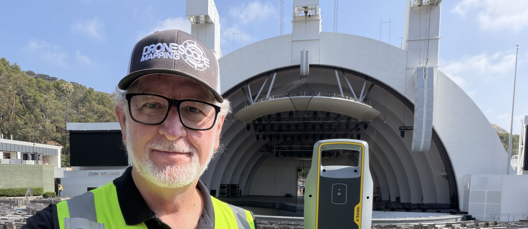 A Drone Scope Mapping operator beside a tripod-mounted laser scanner on the apron of an outdoor amphitheatre, the stage shell behind him.