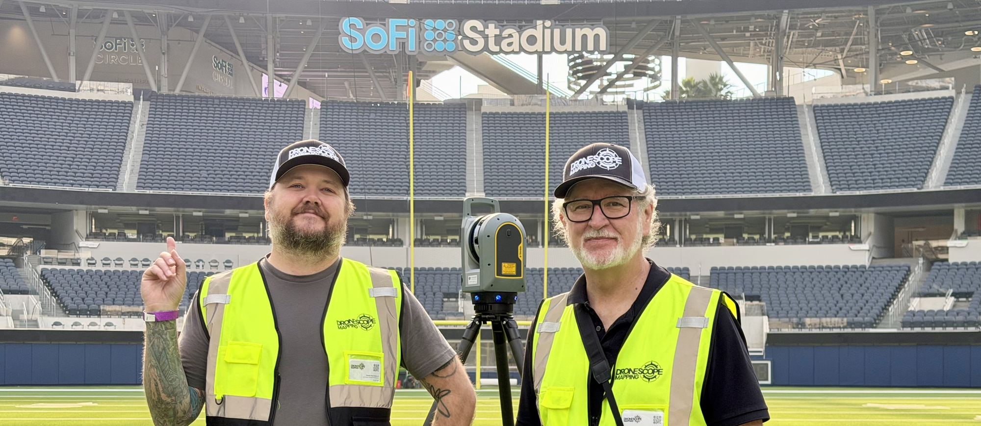 Two Drone Scope Mapping staff in high-visibility vests on the field of an empty stadium, standing either side of a tripod-mounted laser scanner.