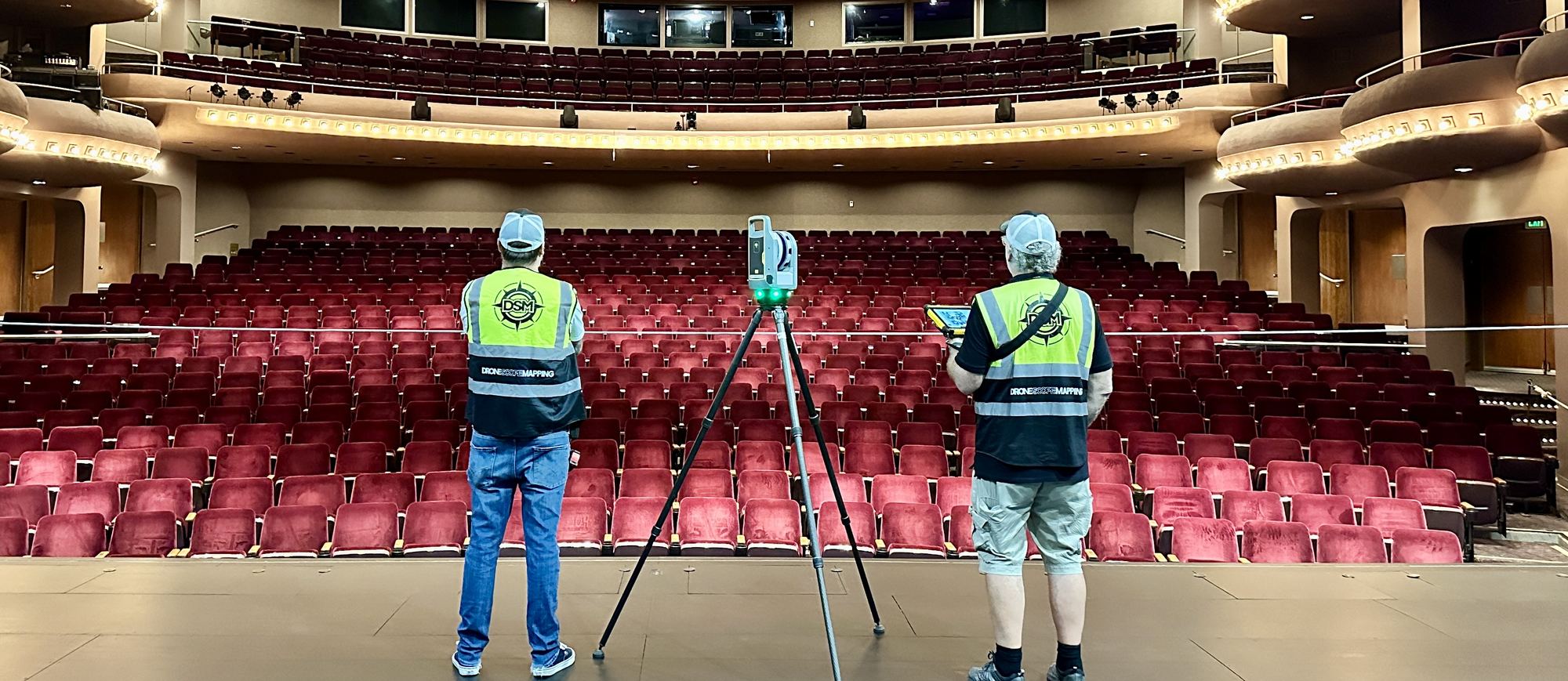 Two Drone Scope Mapping staff either side of a tripod-mounted laser scanner in a theatre auditorium, facing the empty seating and balconies.